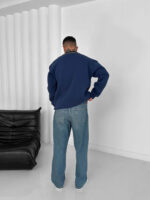 Man wearing oversized navy blue polo sweatshirt with light blue jeans in minimal studio setting.