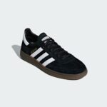 Close-up of a black suede Adidas sneaker with white three stripes and a brown sole, set against a pale gray background.