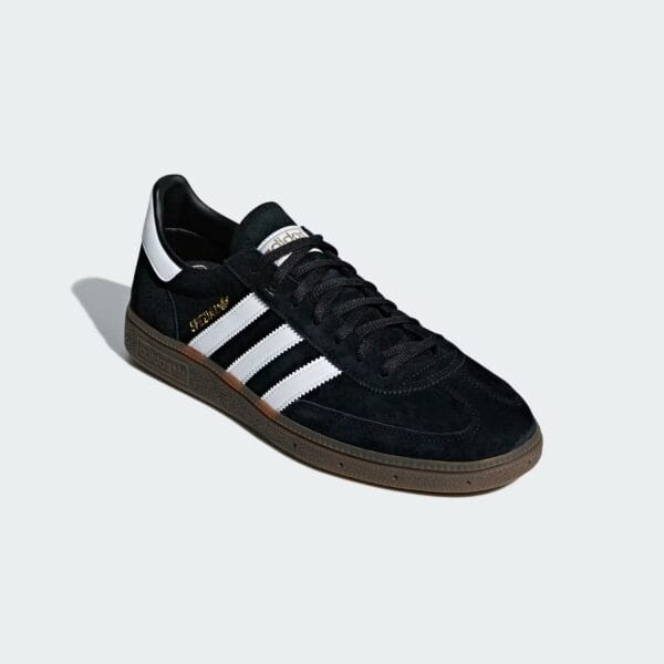 Close-up of a black suede Adidas sneaker with white three stripes and a brown sole, set against a pale gray background.