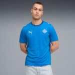 Young man in a bright blue Puma T-shirt with a crest logo, standing with hands behind his back against a gray gradient background.