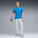 Man wearing a blue athletic shirt and light gray pants stands in a studio, holding a blue and white volleyball against a gray gradient background.