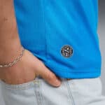 Close-up of a person wearing a bright blue polo with a small circular logo near the hem on the side, hand in jeans pocket.