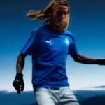 Athlete in a blue Puma jersey mid-action outdoors with a clear blue sky in the background and wind-blown hair.