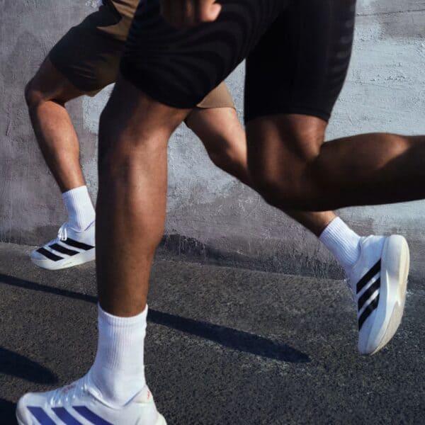 Close-up of two sprinters' legs and white socks, one wearing white Adidas sneakers, on a concrete ground.