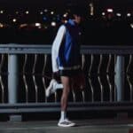 Person in a blue and white athletic jacket standing at a railing on a dark night, city lights blurred in the background.