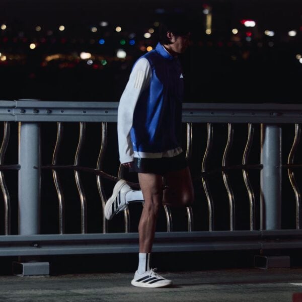 Person in a blue and white athletic jacket standing at a railing on a dark night, city lights blurred in the background.
