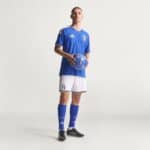Full-body shot of a male soccer player in a blue Italy jersey holding a blue soccer ball, standing against a light neutral background.
