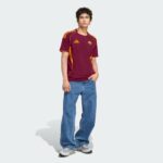 Young man wearing a maroon sports jersey with orange stripes and blue jeans, standing with hands in pockets against a light background.