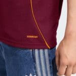Close-up of a maroon AEROREADY sweater with an orange diagonal seam, paired with blue jeans; a hand appears on the right side of the image.
