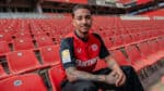 Bayer Leverkusen 2024/25 home football jersey on a model in stadium seats.