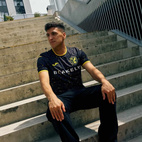 Man wearing Norwich City away football jersey 2024/25 sitting on stairs outdoors.