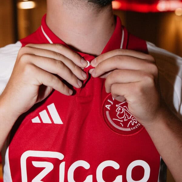 Close-up of person adjusting red AJX HOME football jersey 2024/25 with club logo and Adidas branding.