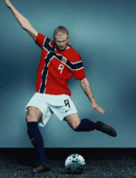 Soccer player in a red jersey with a crest, white shorts and blue socks, mid‑kick toward a white soccer ball on a dark surface.