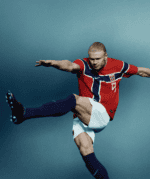 Male soccer player in a red jersey with a Norwegian flag design, mid-air kick against a blue backdrop.