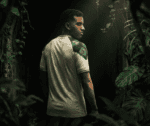 Man in a light polo stands in dense jungle, turned slightly to the side, with a green tropical leaf pattern on his shoulder.