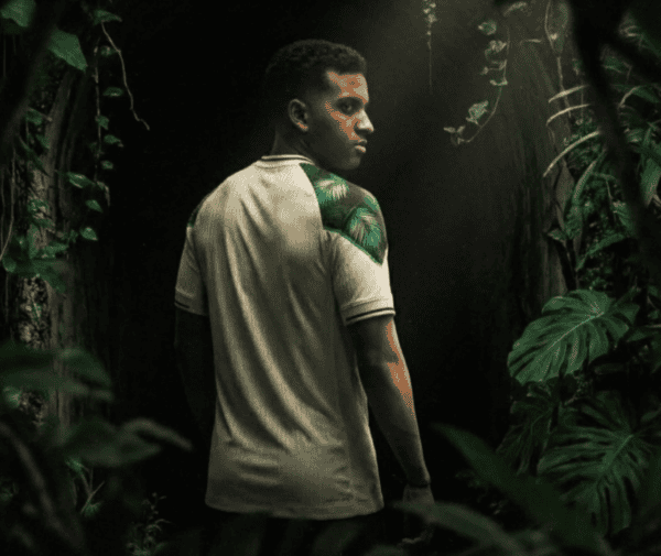 Man in a light polo stands in dense jungle, turned slightly to the side, with a green tropical leaf pattern on his shoulder.