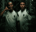 Two men in Brazil national team shirts stand among dense tropical plants in a dim jungle setting.