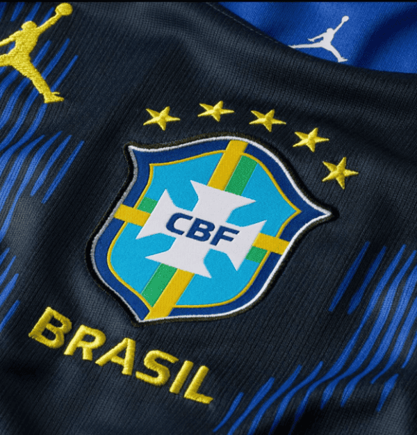 Close-up of Brazil national football jersey showing the CBF crest, four gold stars, and 'BRASIL' text on dark fabric with blue and green accents.