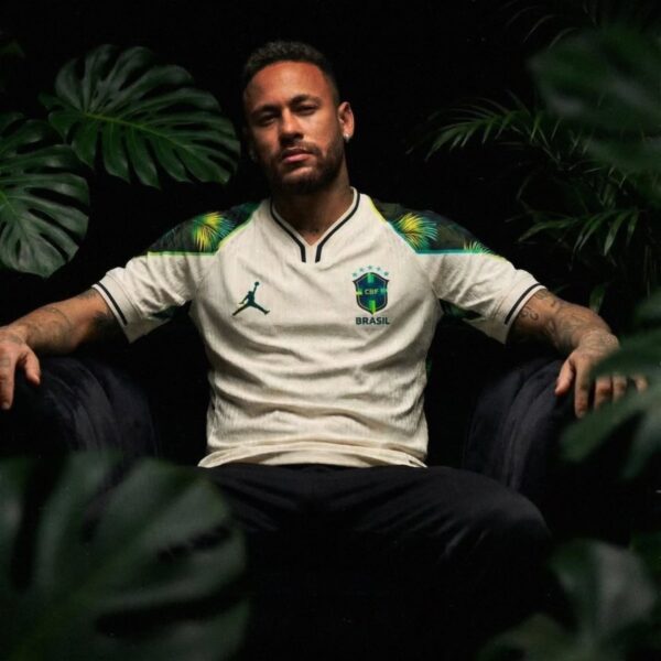 Man seated in a dark setting wearing a white Brazil national team jersey, arms resting on the chair arms.