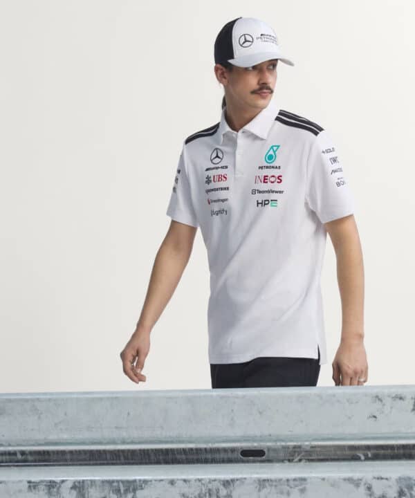 Man in a white polo with sponsor logos and a Mercedes cap standing by a metal railing, looking to the side.