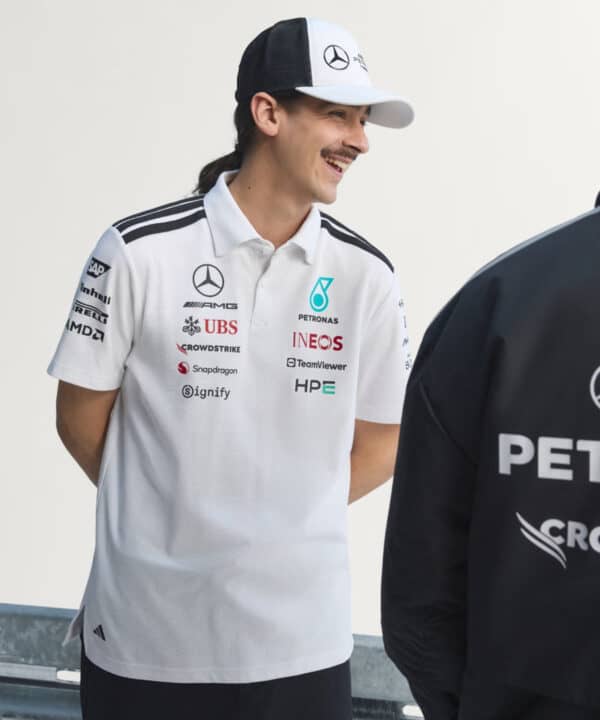 Smiling man in a white polo with multiple sponsor logos and a white cap (Mercedes) facing to the right.