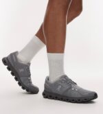 Gray On running shoes with a chunky black sole, worn with light gray crew socks in a mid-stride pose.