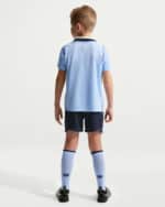 Back view of a young soccer player wearing a light blue jersey, dark shorts, white knee-high socks, and cleats.
