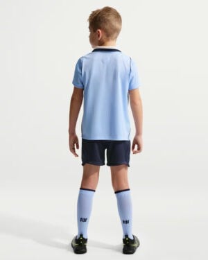 Back view of a young soccer player wearing a light blue jersey, dark shorts, white knee-high socks, and cleats.