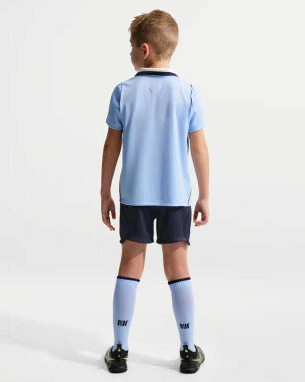 Back view of a young soccer player wearing a light blue jersey, dark shorts, white knee-high socks, and cleats.