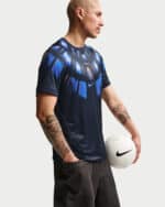 Male model in a dark blue patterned soccer jersey, holding a white Nike soccer ball, seen in profile with tattoos on both arms.