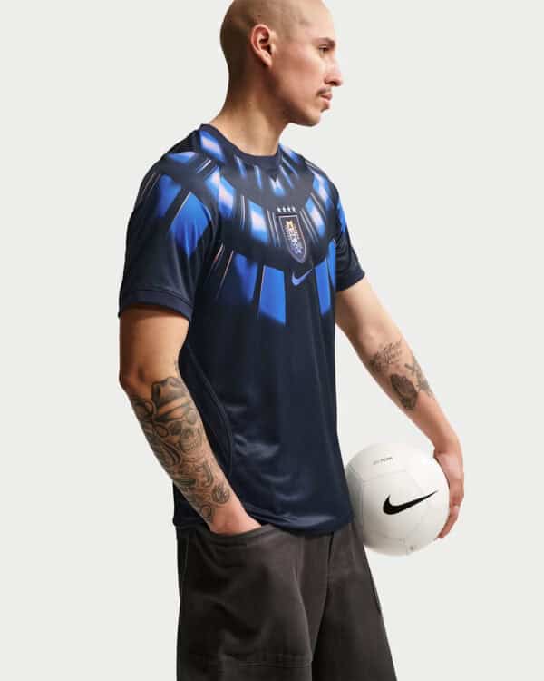 Male model in a dark blue patterned soccer jersey, holding a white Nike soccer ball, seen in profile with tattoos on both arms.