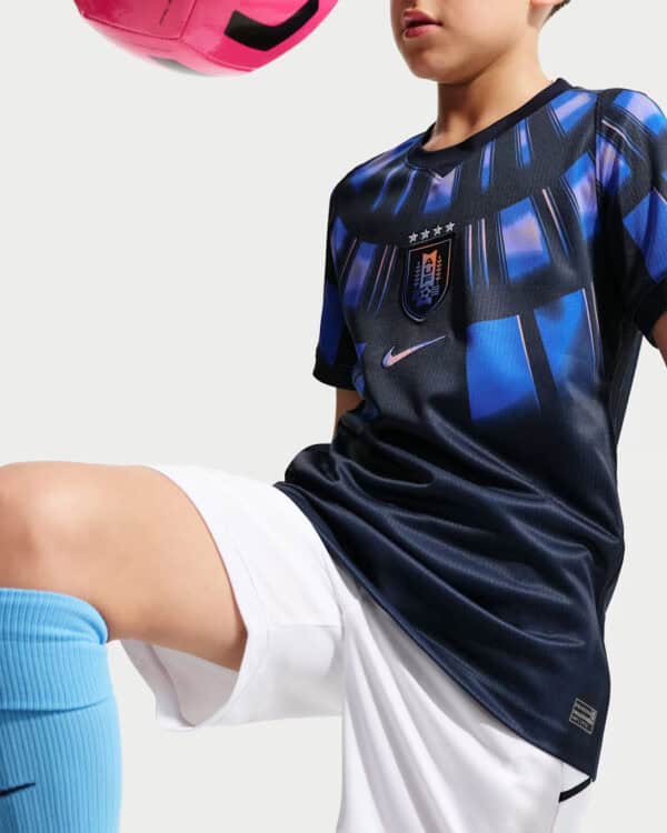 Child wearing a dark blue soccer jersey with a USA crest, white shorts, and blue socks, kicking a pink ball partially visible at the top corner.