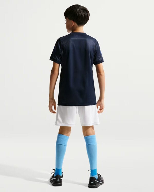 Back view of a young boy wearing a navy soccer jersey, white shorts, bright blue knee-high socks, and black cleats.