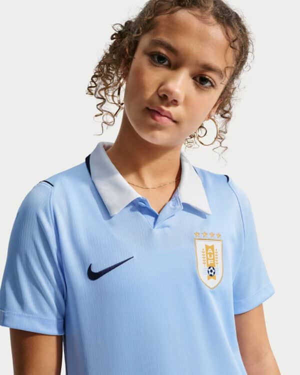 Young girl wearing a light blue soccer jersey with a crest and Nike logo, looking at the camera.