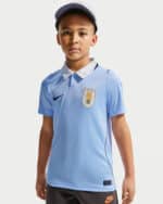 Boy wearing a light blue soccer jersey with a crest and cap, facing the camera against a plain background,