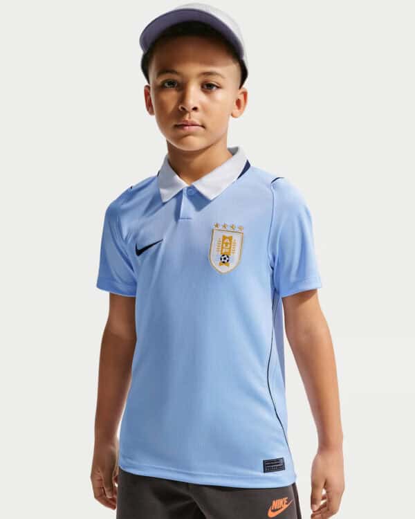 Boy wearing a light blue soccer jersey with a crest and cap, facing the camera against a plain background,