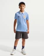 Young boy modeling a light-blue polo soccer jersey with a crest, paired with dark shorts and sneakers.