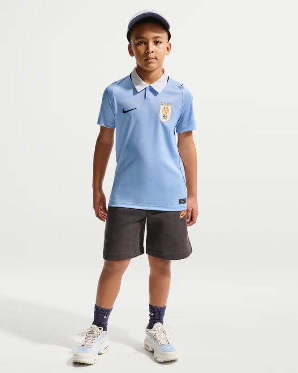 Young boy modeling a light-blue polo soccer jersey with a crest, paired with dark shorts and sneakers.