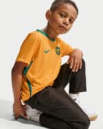 Young boy in an orange sports jersey with green trim kneeling on the floor, facing the camera.