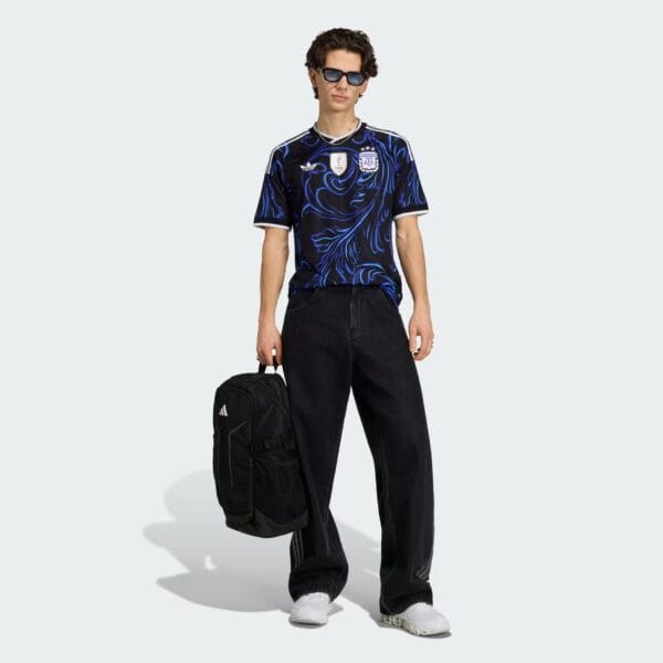 Male model wearing a black-blue swirling graphic jersey, sunglasses, dark jeans, white sneakers, and carrying a black backpack