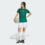 Female soccer player in a green jersey holding a multicolored soccer ball against a light background, facing left with hands on hips.