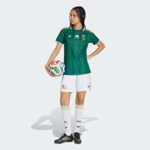 Female soccer player in a green jersey holding a multicolored soccer ball against a light background, facing left with hands on hips.