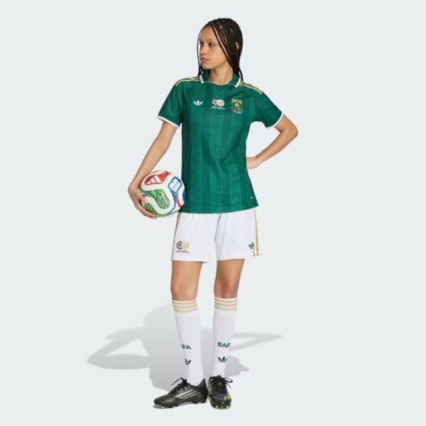 Female soccer player in a green jersey holding a multicolored soccer ball against a light background, facing left with hands on hips.