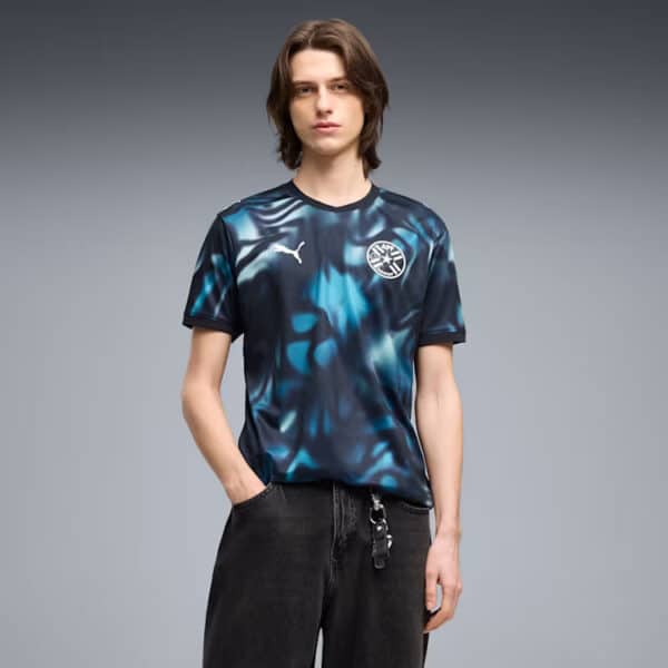Young man wearing a blue abstract-patterned soccer jersey with a white Puma logo and a circular club crest, standing with hands in pockets.