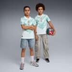 Two kids wearing matching Portugal soccer jerseys stand side by side, one with arms crossed and the other holding a turquoise ball under his arm.