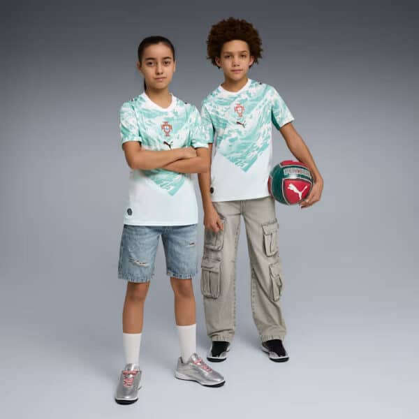 Two kids wearing matching Portugal soccer jerseys stand side by side, one with arms crossed and the other holding a turquoise ball under his arm.