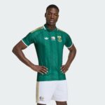 Young man in a green striped football jersey with gold stripes, standing with hands on hips, white shorts, and SA Football Association logo visible