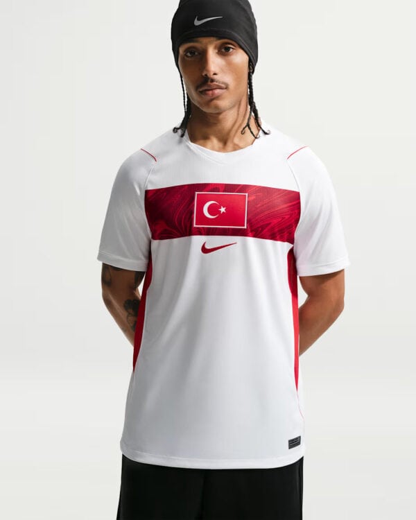 Male model wearing a white Nike athletic jersey with a red chest band and Turkish flag, against a light background, wearing a black beanie.