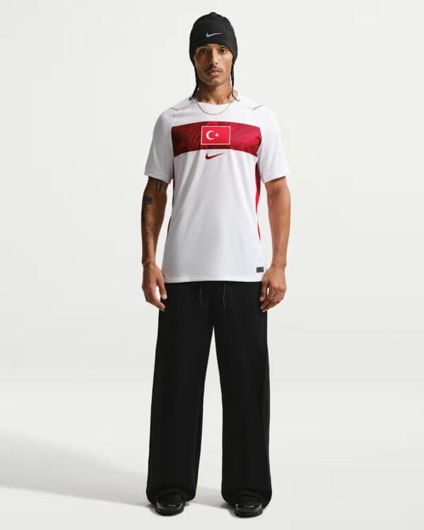 Male model wearing a white athletic shirt with a red Turkish flag stripe across the chest, black wide-leg pants, and a black beanie.