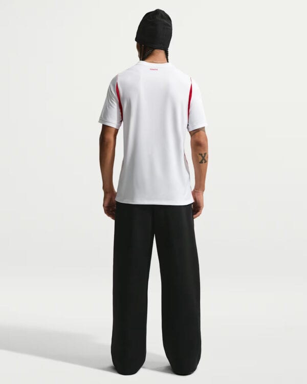 Person standing with back to the camera, wearing a white short-sleeve shirt with red side panels, wide black pants, and a black beanie.